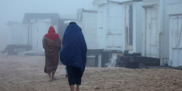 Two people, wrapped in sleeping bags, walk along the beach among makeshift huts