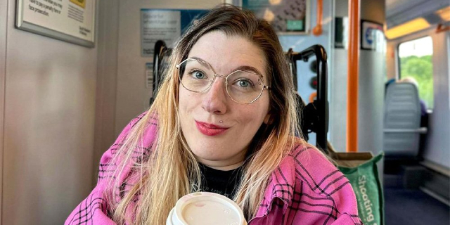 Melody Powell on a train wearing a pink coat and holding a Starbucks coffee cup. She is looking into the camera and smiling