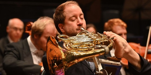 French horn player Felix Klieser is pictured playing in the Bournemouth Symphony Orchestra. The disabled musician plays his instrument with his feet as he was born without arms. He is dressed in a suit and tie and other musicians can be seen in the background