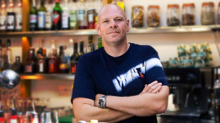 TV chef Tom Kerridge stands looking into camera with his arms folded. He is wearing a navy blue t-shirt and is standing in a kitchen with various jars and bottles behind him