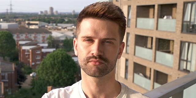 Sam, a case study from a BBC story about renters who were unable to access affordable housing schemes. He is wearing a white t-shirt and standing on the balcony of a London flat while looking into the camera