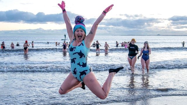 A woman wearing a swimming costume and a woolly hat does a starjump as dozens of other women swim in the sea behind her during a sunrise dip in the Firth of Forth at Portobello beach in Edinburgh on International Women's Day 2023