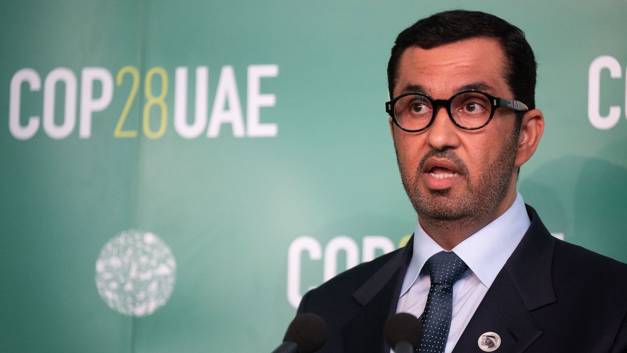 COP28 President Sultan al-Jaber wears a suit and tie and stands in front of a sign for the conference