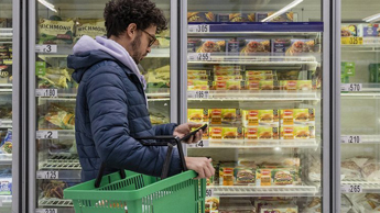 A man checks his phone, while carrying a basket in a supermarket freezer aisle